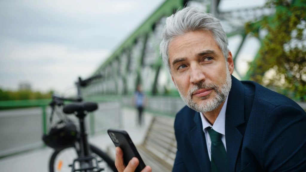 A bearded man in a suit sitting on a bench and looking to the side while holding his phone.
