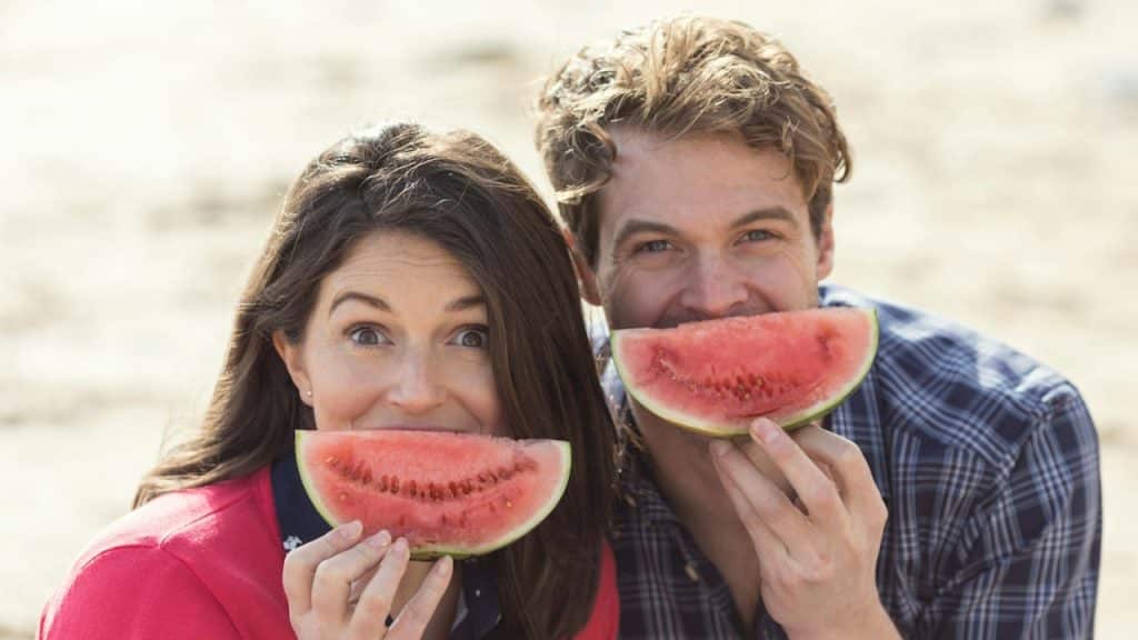 A couple holding watermelon slices up to their mouths and being happy.