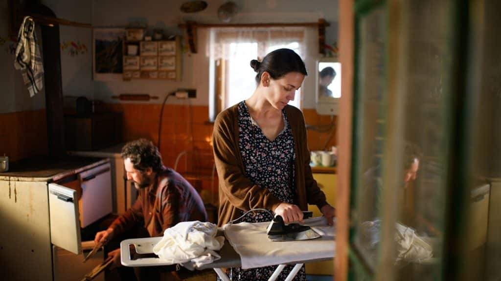 A woman ironing clothes in a dimly lit, rustic kitchen while a man fixes wooden cabinets in the background.