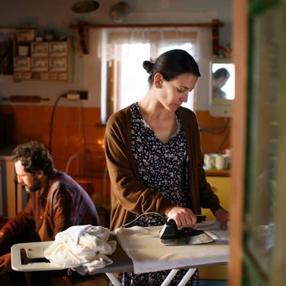 A woman ironing clothes in a dimly lit, rustic kitchen while a man fixes wooden cabinets in the background.