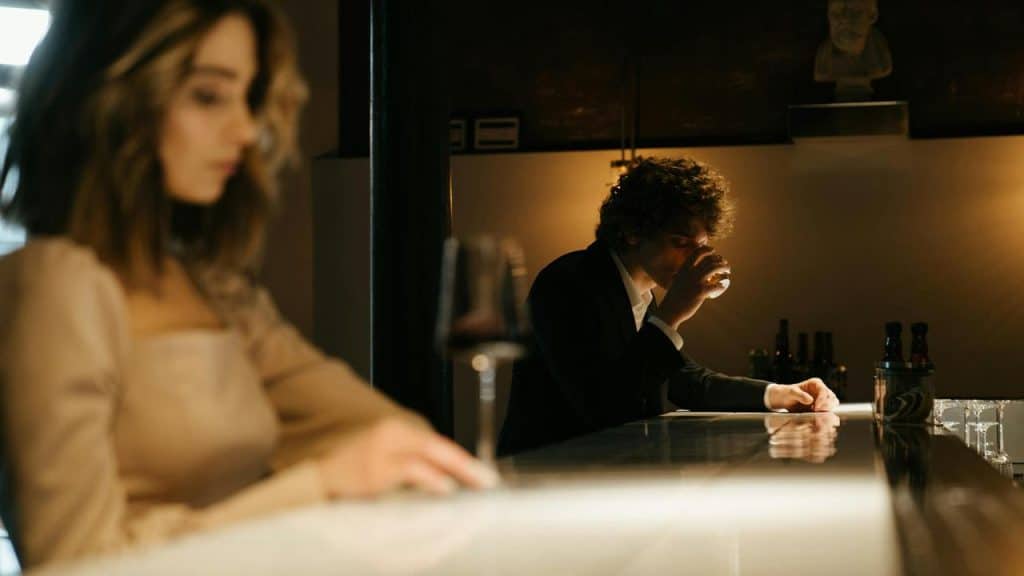A man drinking alone at a bar while a woman sits nearby.