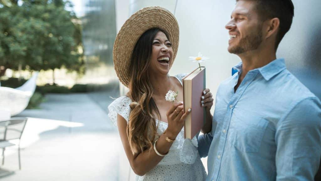 A woman laughing and holding a book while standing with a smiling man outdoors.