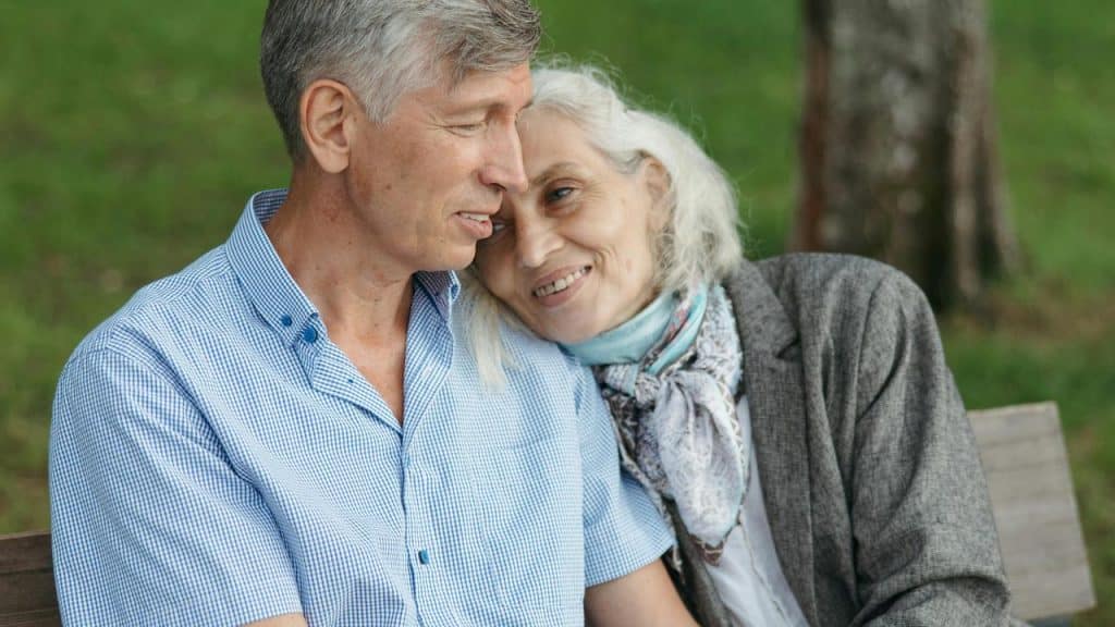 An elderly couple sitting close together on a bench and smiling.
