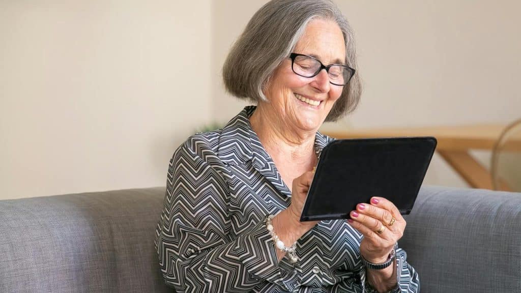 An elderly woman smiling while using a tablet on a couch.