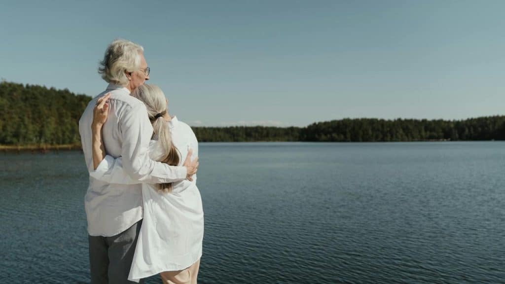 An elderly couple embracing while looking out over a lake.