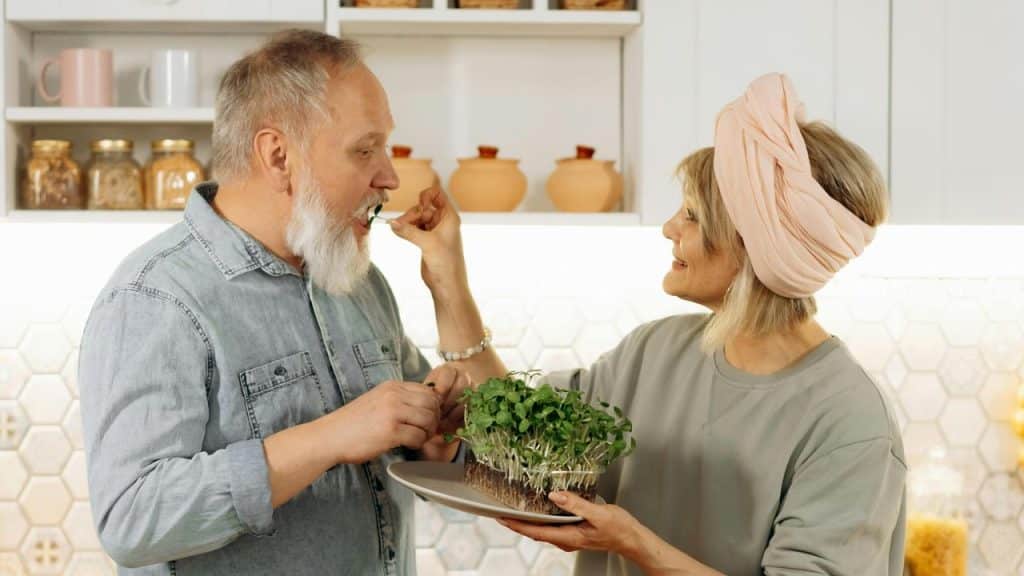 An older woman feeding a man fresh greens in a kitchen.