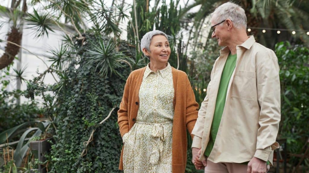 An elderly couple holding hands and smiling while walking through a garden.