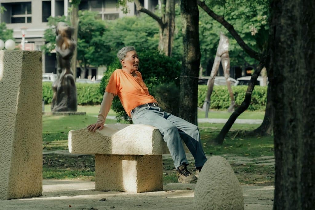 The relaxed man in an orange shirt is sitting at the park.