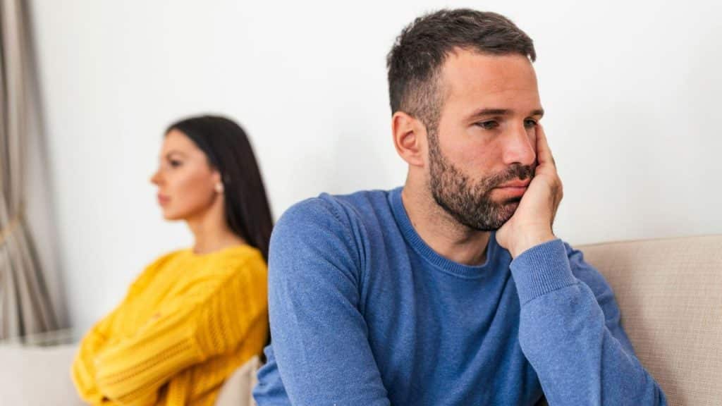 Man in blue sits looking sad, chin resting on his hand, while a woman in yellow sits with her back to him.