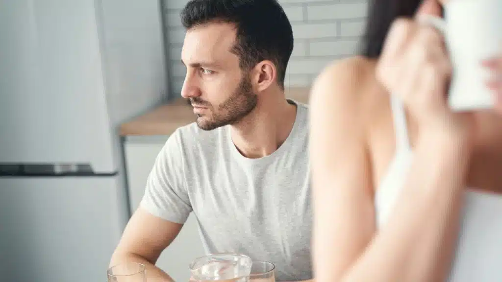 A bearded man in a gray shirt sits at a table in a kitchen, looking away with a serious expression.