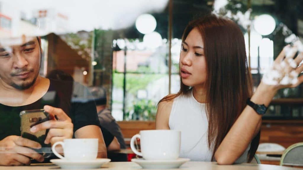 A man looks at his phone while a woman across from him gestures and speaks with a frustrated expression.