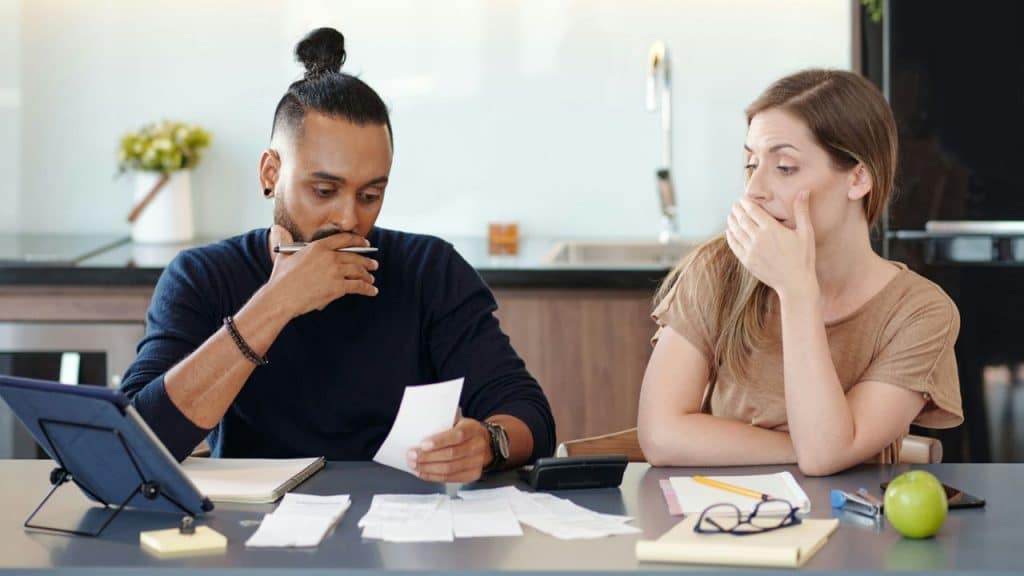 A man with a topknot looks intently at a receipt while a woman next to him holds her hand over her mouth.