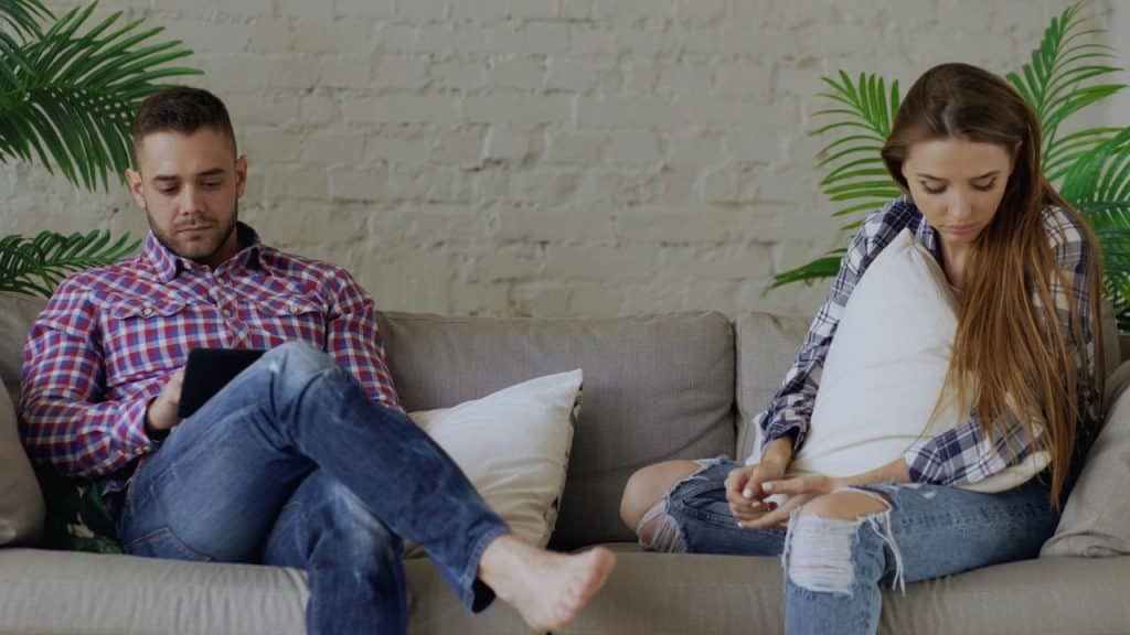 A man looks at a tablet while a woman sits sadly away from him, holding a pillow.