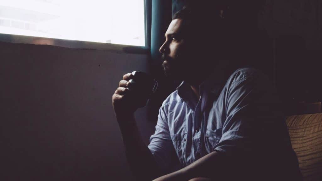A bearded man sits in a dark room, holding a mug and looking out a brightly lit window.