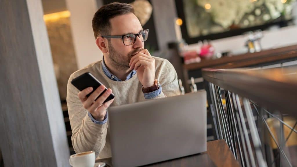 A man in glasses sits at a table with a laptop, holding a phone and looking away thoughtfully.
