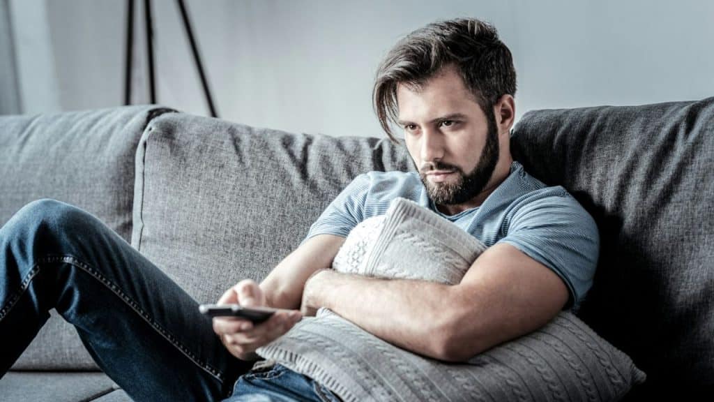 A bearded man sits on a gray couch, holding a remote and a pillow, looking intently at a screen.