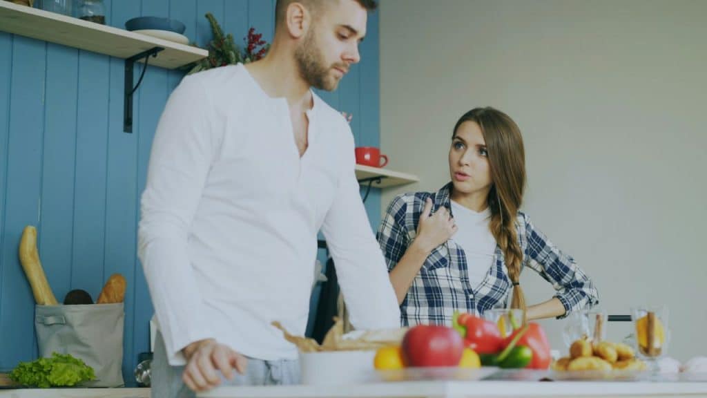 A man stands ignoring a woman in a plaid shirt who is gesturing and speaking to him in a kitchen.
