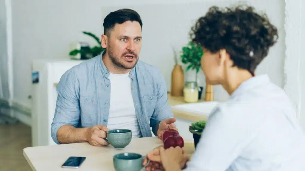A man sits at a table with a mug, speaking to a woman across from him with a surprised expression.