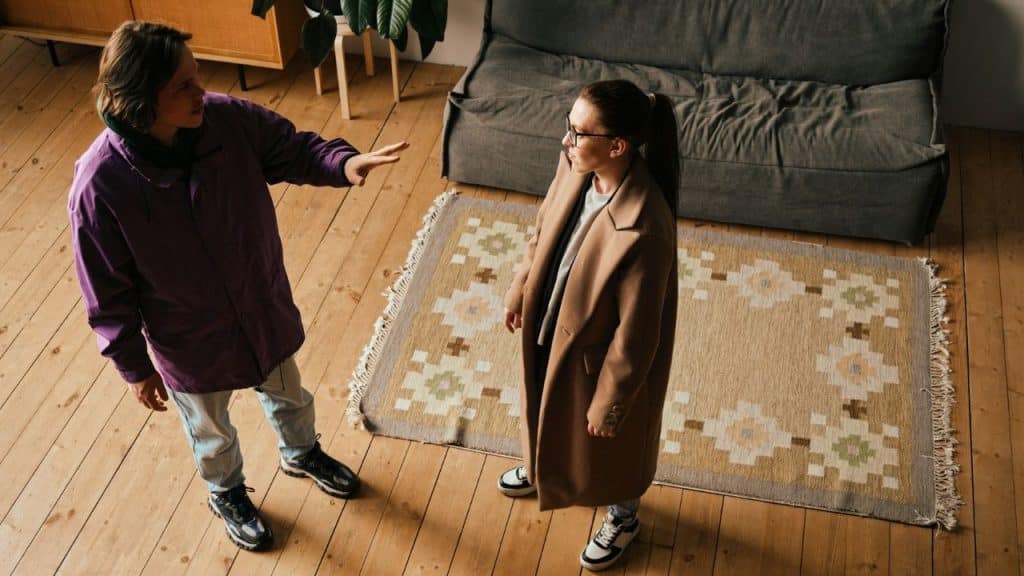Overhead shot of two people in coats arguing in a living room, one gesturing emphatically.