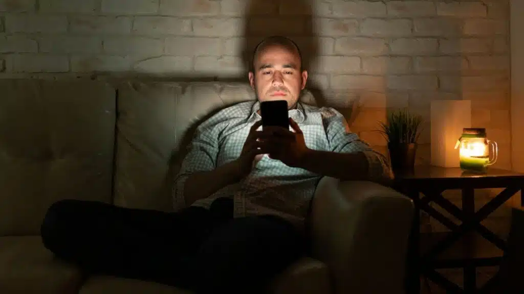 A man sits alone on a couch in a dim room, looking down at his phone illuminated by its screen.