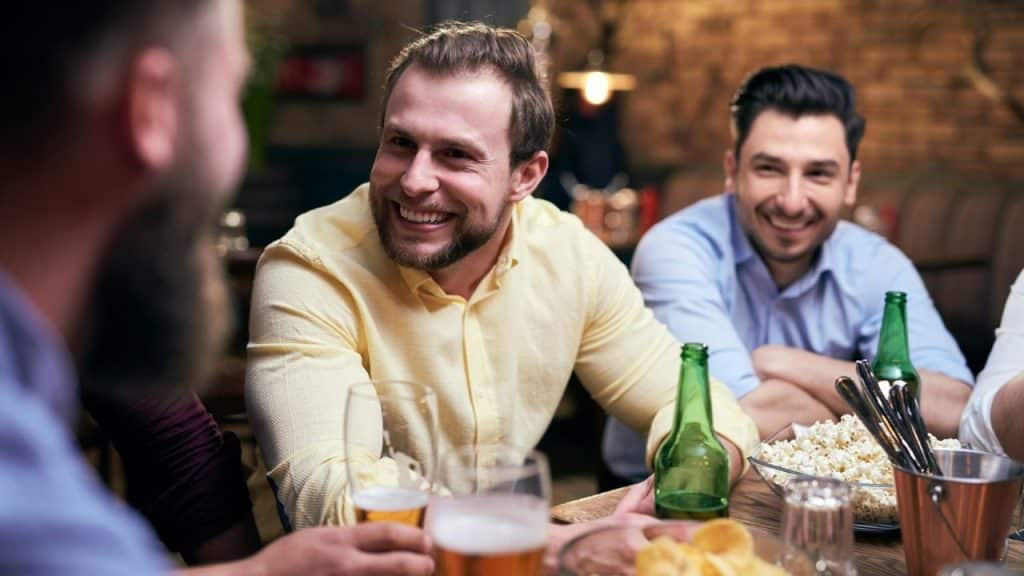 Three bearded men sit around a table with beer and snacks, smiling and talking in a dimly lit bar.