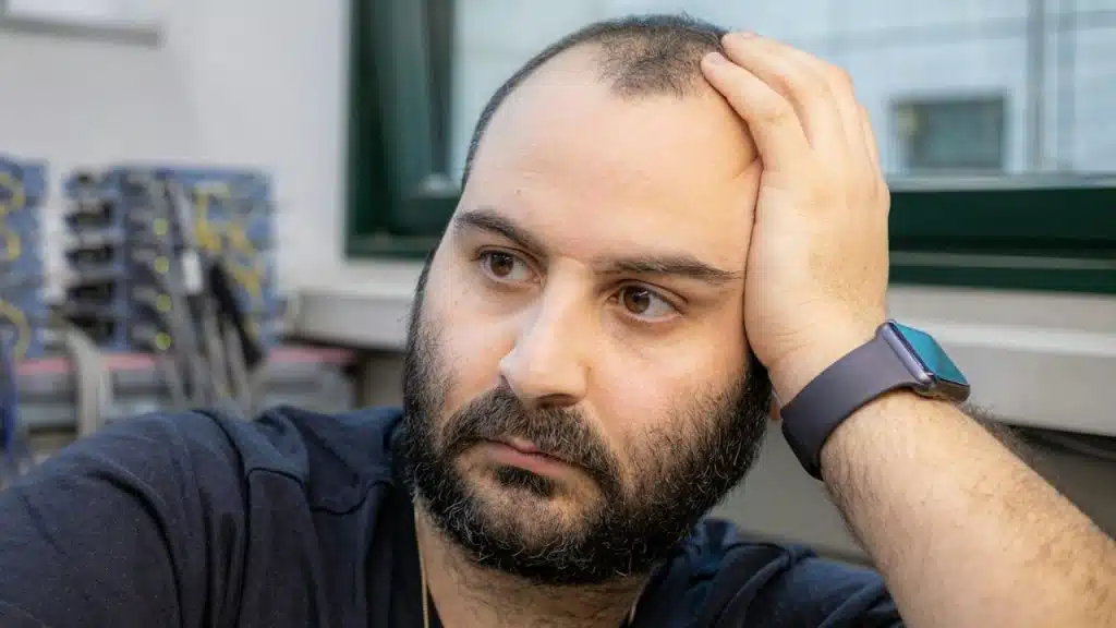 Man with a beard rests his hand on his head, looking off-camera.