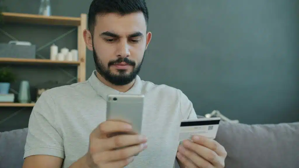 A bearded man in a light shirt is looking at his phone while holding a credit card.