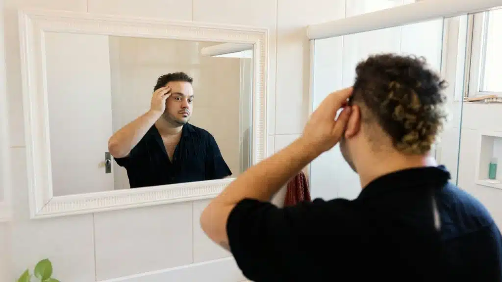 A man looks into a bathroom mirror, touching the top of his head with one hand.