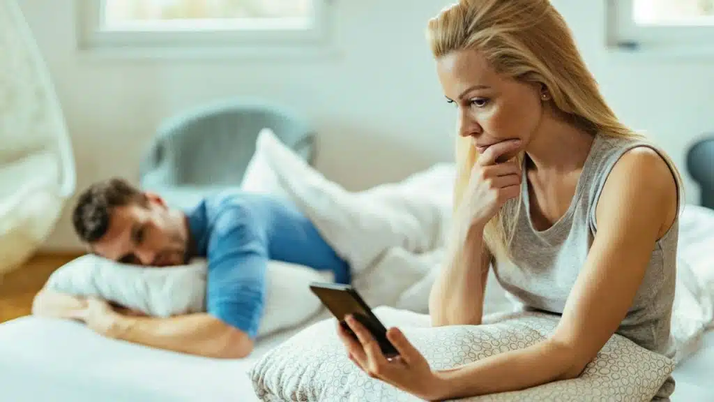 A woman sits up in bed looking at her phone while a man lies next to her.