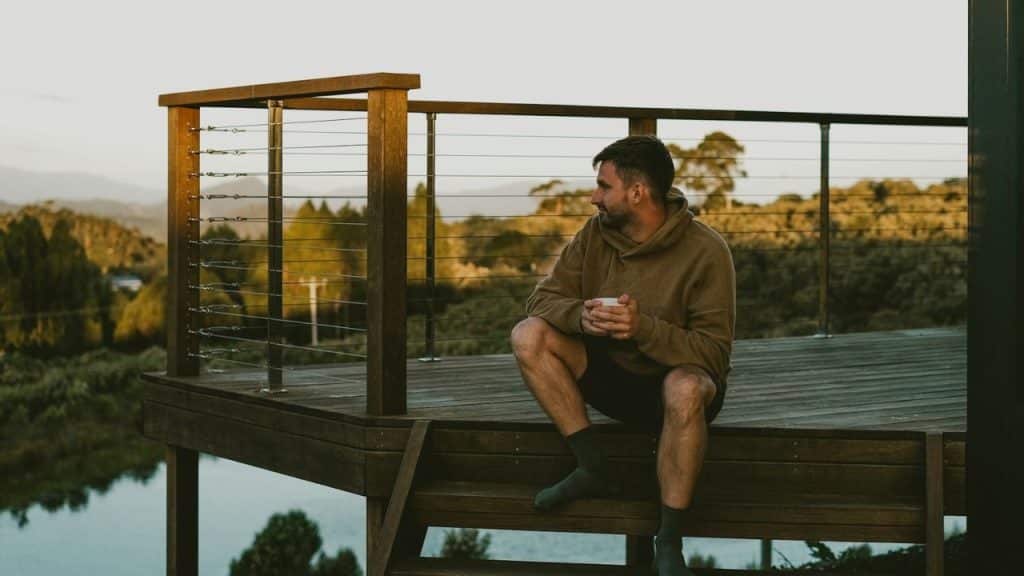 A man sits on deck steps, holding a cup and looking out at a hilly landscape.
