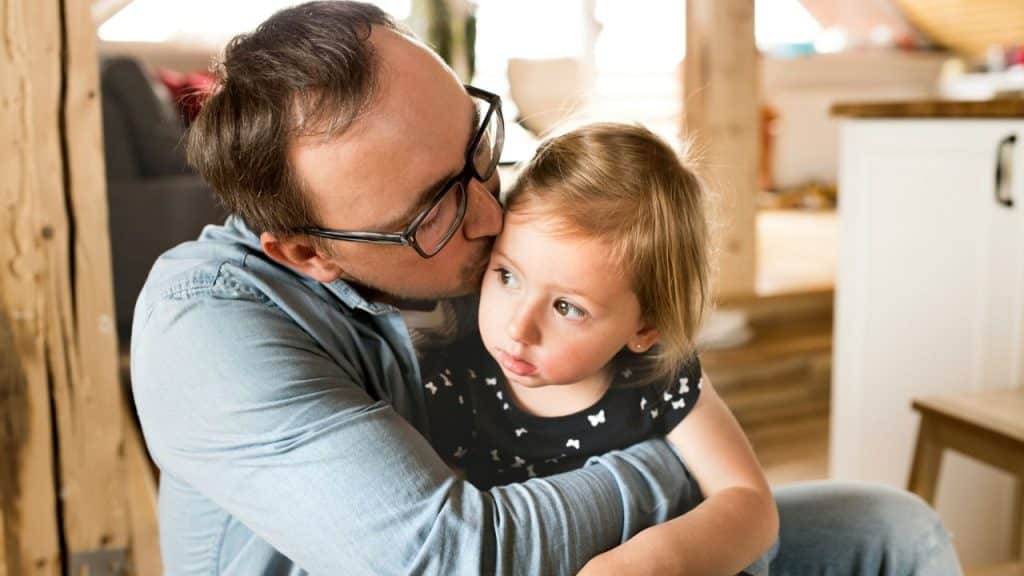 A man with glasses is hugging a young girl and kissing her on the cheek.