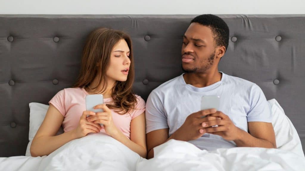 A couple is in bed, both looking at their phones, but the man is looking sideways at the woman.