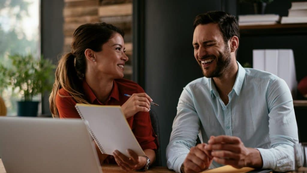 A man and woman are sitting at a desk, smiling at each other during a conversation.