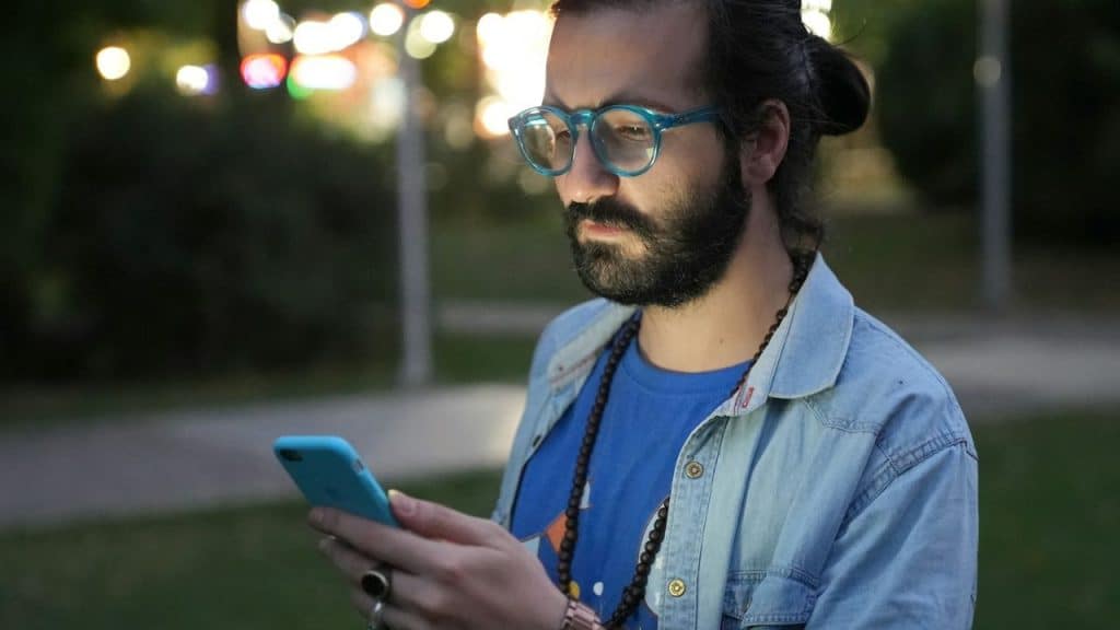 A bearded man in a denim shirt and blue glasses looks down at his phone outdoors at night.