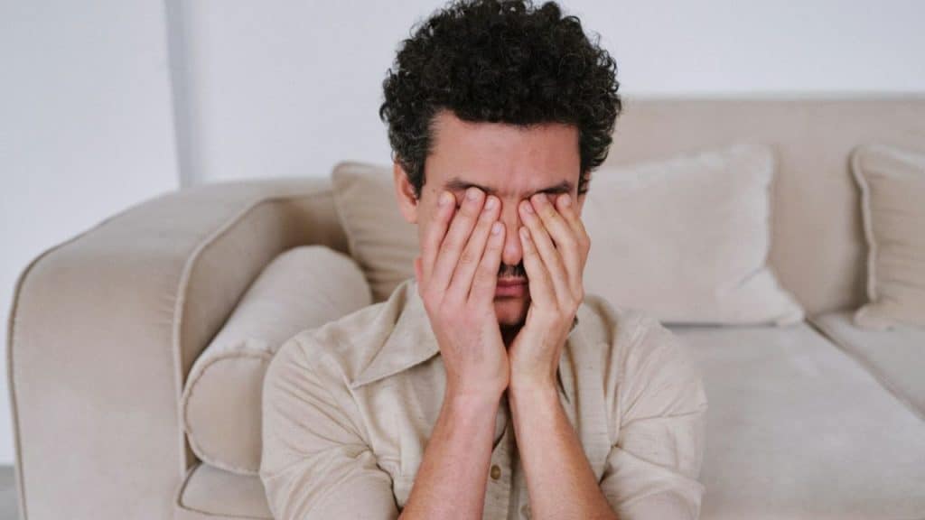 A man with curly hair sits on the floor, covering his face with both hands.