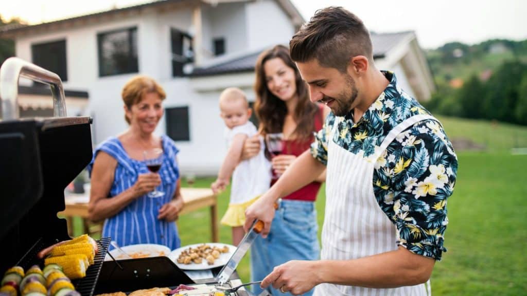 A man is grilling outdoors while a woman, a baby, and an older woman stand behind him.