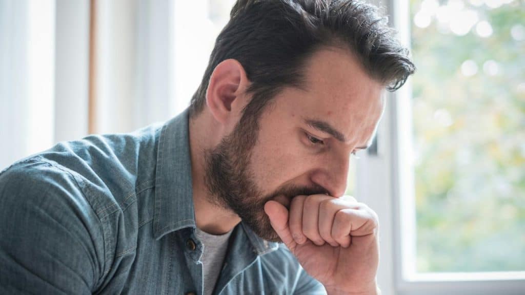 A bearded man in a denim shirt looks down thoughtfully, resting his fist on his mouth.