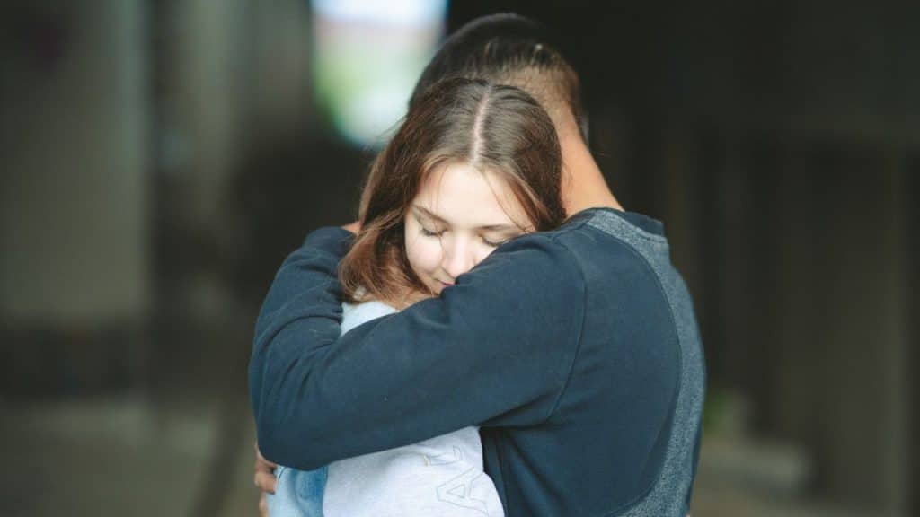 A man in a dark shirt is hugging a woman with long hair, who has her eyes closed.