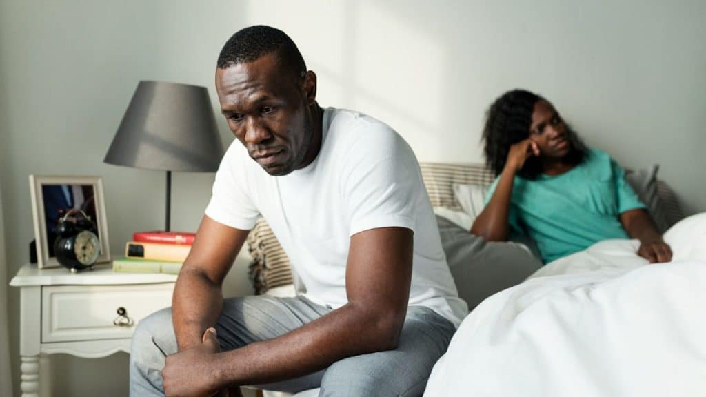 A man sits on the edge of a bed looking down sadly, while a woman sits behind him looking away.