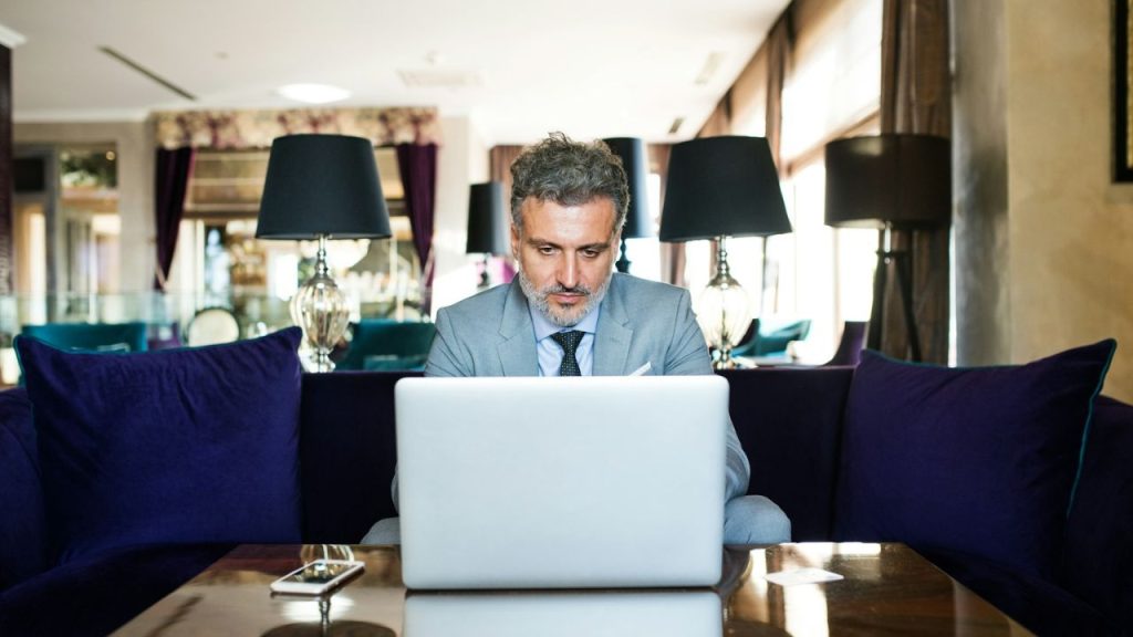 A man in a suit is sitting on a couch, looking intently at a laptop in front of him.