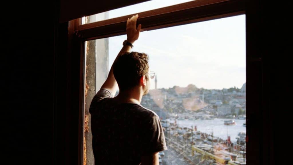 A man with glasses stands facing a window with his hand up, looking out over a city and harbor.