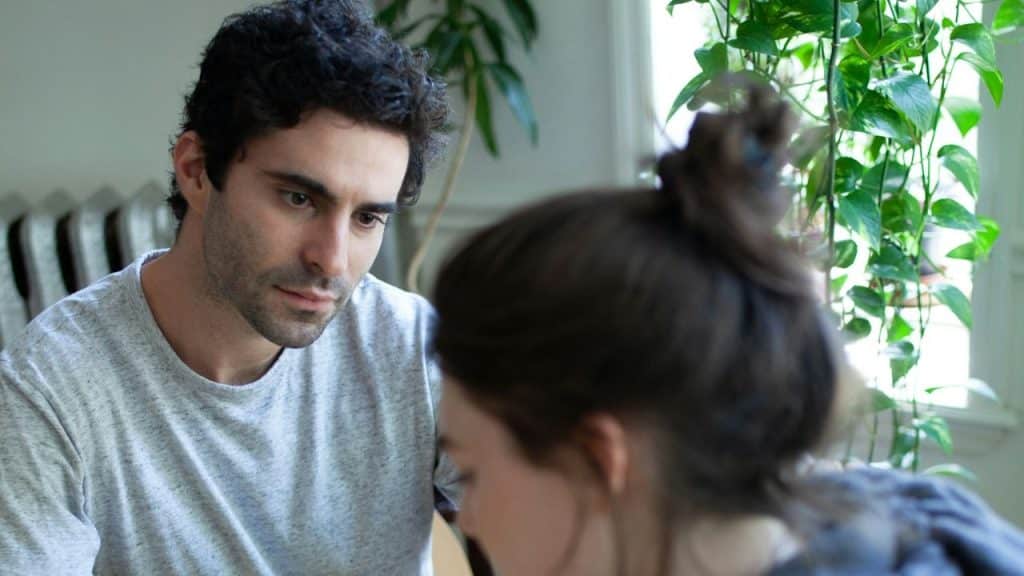 A man with dark, curly hair looks down with a concerned expression at a person out of focus in the foreground.