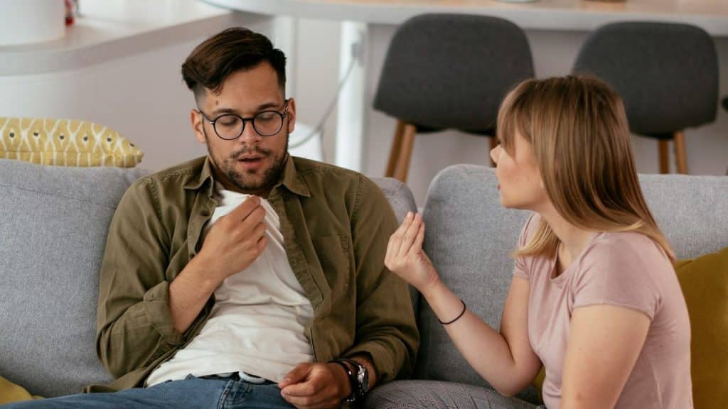 A woman is sitting next to a man on a couch, speaking and gesturing toward him as he looks down.