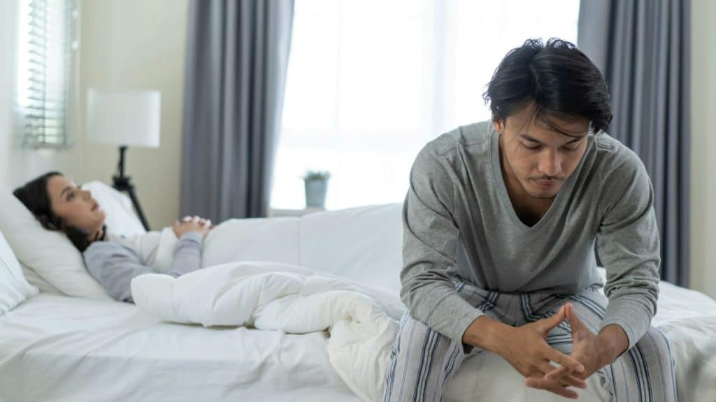 A man sits on the edge of the bed looking down while a woman lies under the covers in the background.