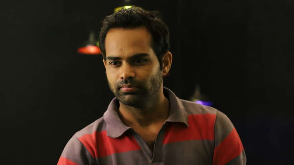 A man in a striped polo shirt is looking down with a serious expression in a dimly lit setting.