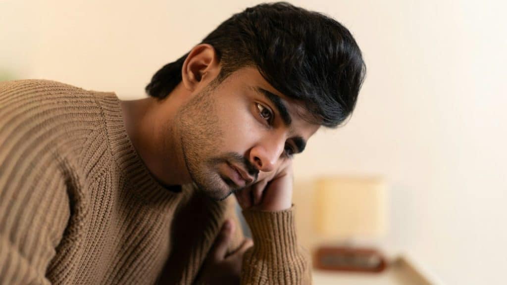 A young man in a brown sweater is sitting with his head resting on his hand, looking down sadly.
