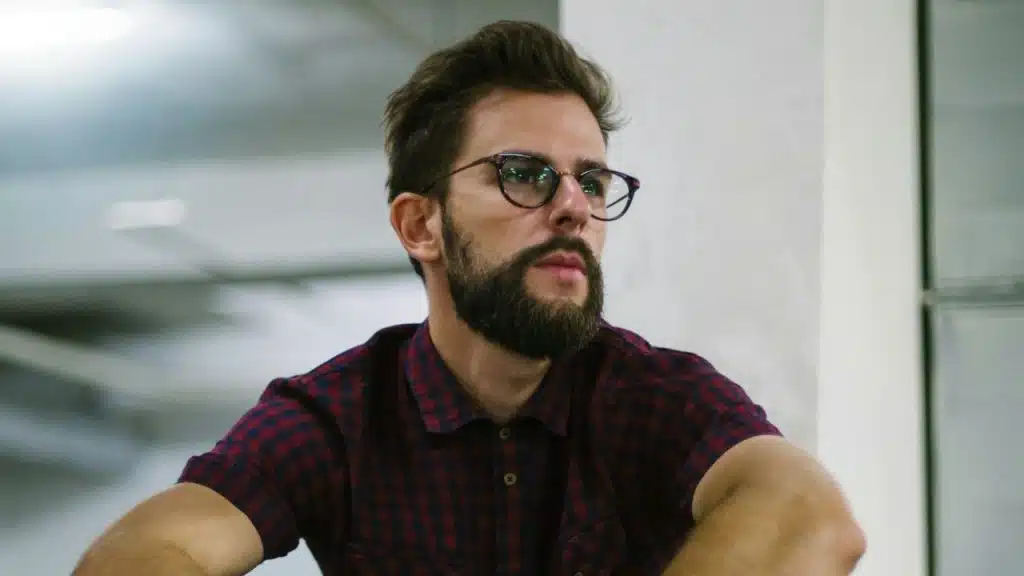 A bearded man with glasses and a plaid shirt is looking off-camera.