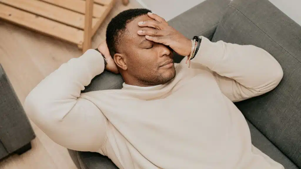 A man is lying on a gray couch with his eyes closed, holding his hands to his forehead and head.