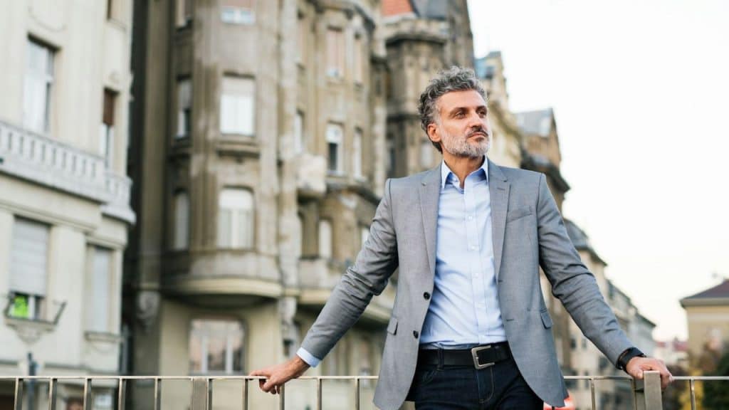A well-dressed, middle-aged man stands leaning on a railing against a backdrop of old city buildings.