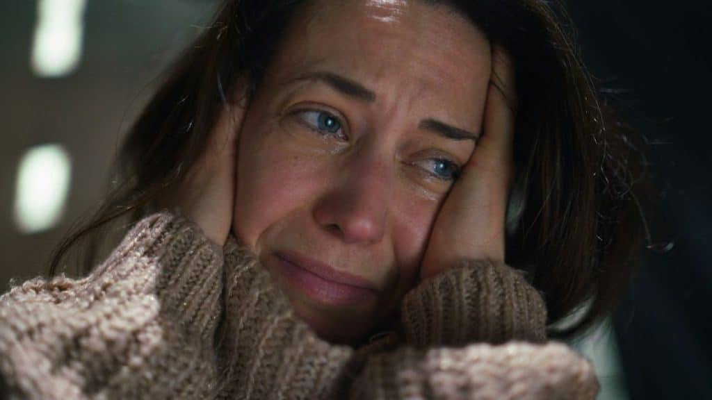 A woman is crying with blue eyes, holding her head with both hands, wearing a brown knit sweater.